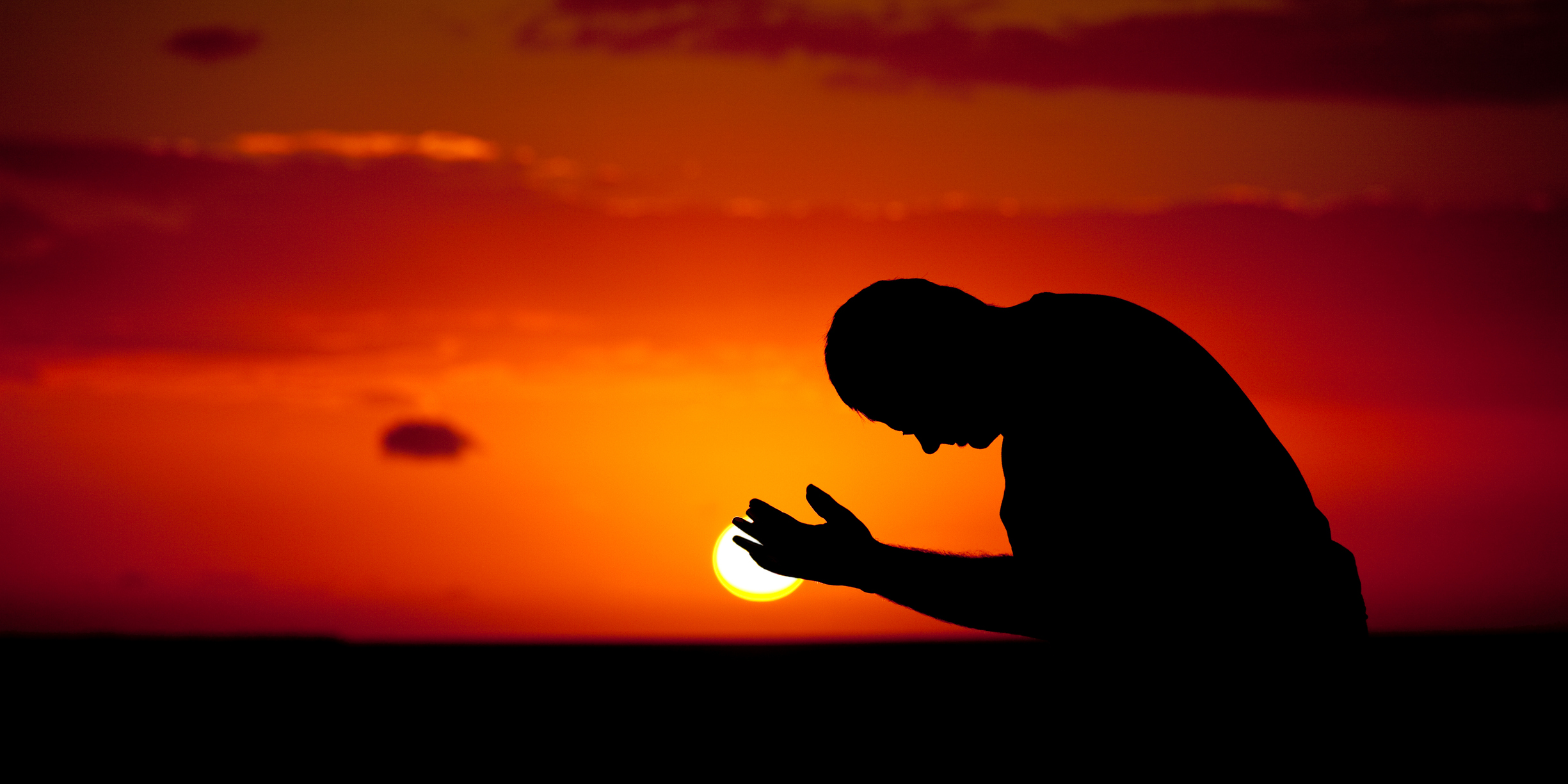 A man praying with hands open. Silhouette. Panoramic image of side view of Caucasian male praying with palms upward. Man is bowing down with ocean and dramatic sun behind. One person is in the image, taking in a tropical climate. Themes include spirituality, balance, lifestyle, salvation, praying, faith, meditation, contentment, vitality, wellness, hope, heaven, healing, deliverance, sin, struggle, help, and relationships.